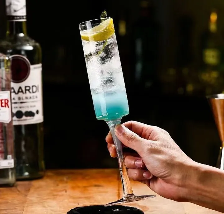 Hand, die ein Cocktailglas mit Stiel hält, gefüllt mit einem Getränk mit blauem Farbverlauf und einer Zitronenscheibe als Garnitur