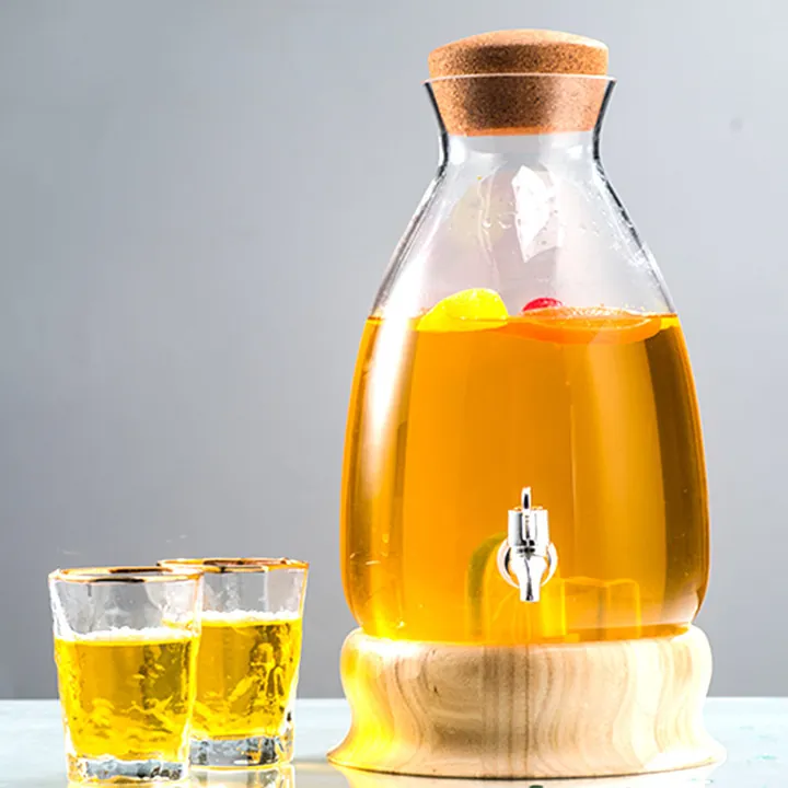 Distributeur de boissons en verre posé sur un socle en bois, contenant une boisson à l'orange, à côté de deux verres
