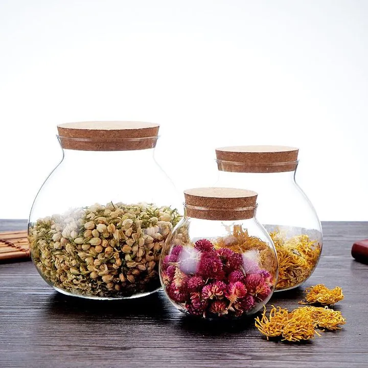 Drei runde Glasbehälter mit Korkdeckeln, gefüllt mit getrockneten Blumen, auf einem Tisch
