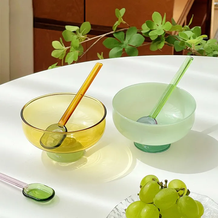 Bernsteinfarbene und hellgrüne Glasschalen mit einem Glaslöffel und Weintrauben auf dem Tisch