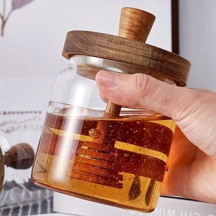 Eine Hand, die ein durchsichtiges Glas mit Honig, einem Holzdeckel und einem Honiglöffel hält