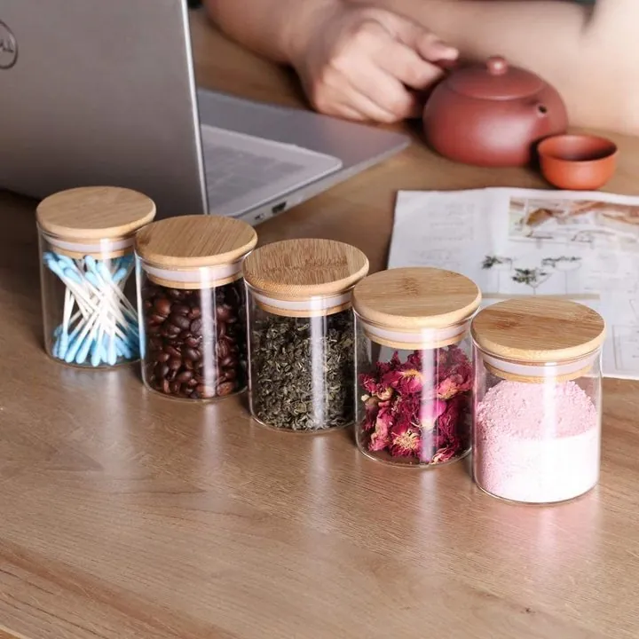 Cinq bocaux de conservation en verre transparent avec des couvercles en bambou, posés sur une table