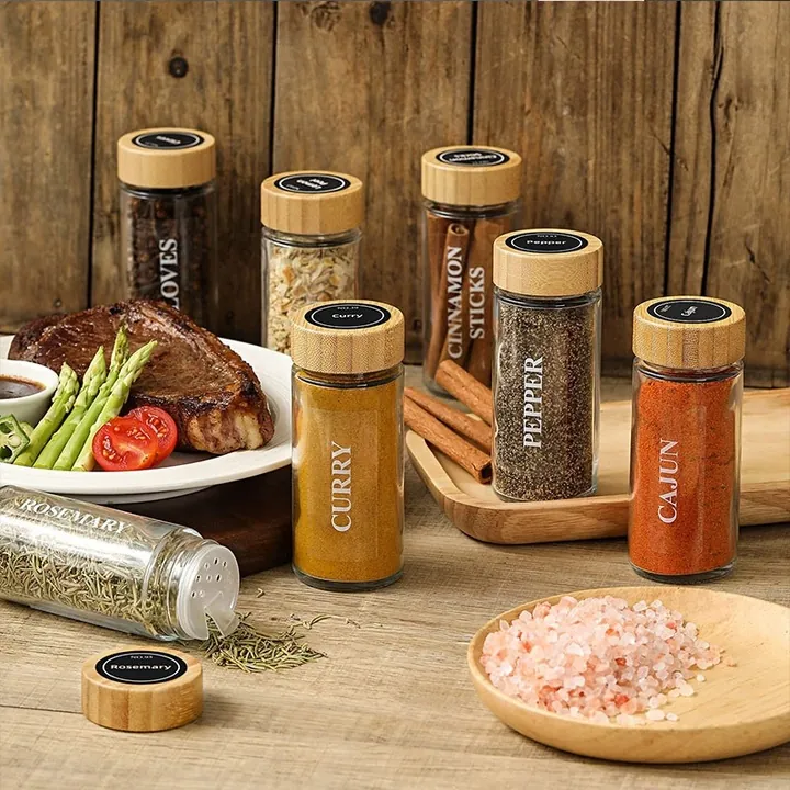 Tarros de cristal para especias con tapa de bambú sobre una mesa de comedor, junto a un filete y condimentos